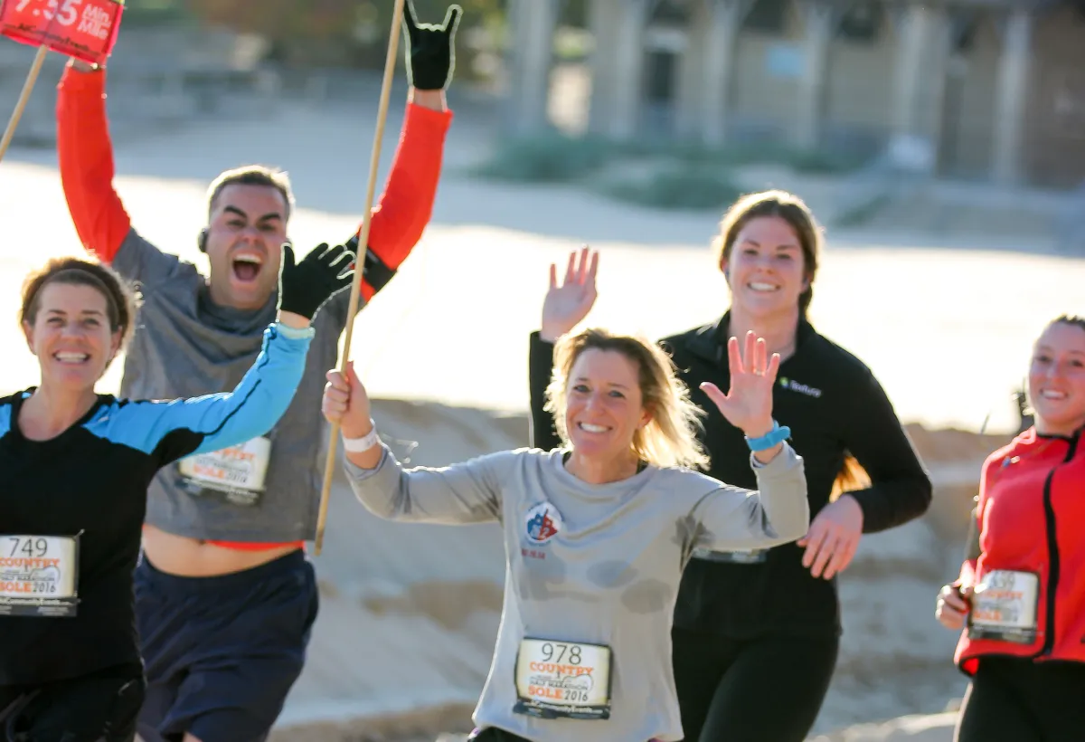 A group of runners smiling and waving, with one runner in the background holding up a sign and making a rock-on gesture, indicating excitement and celebration at a race.