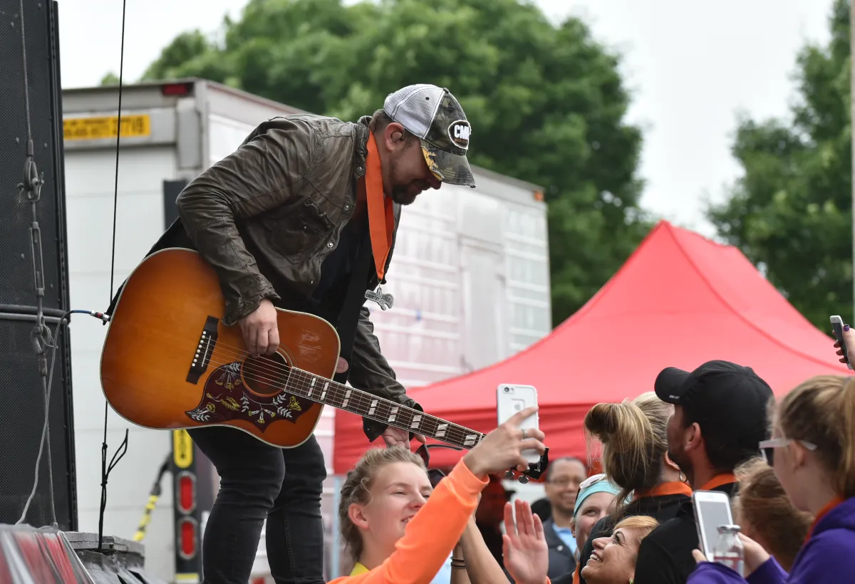 A musician plays guitar on a stage while a crowd of people, some wearing race bibs, watch and take photos with their phones.