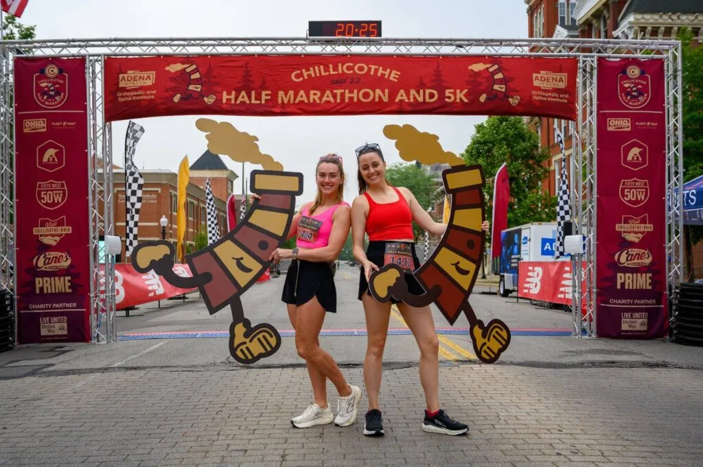 Two women smiling and holding large cartoon cutouts at the finish line of the Chillicothe Half Marathon and 5K.