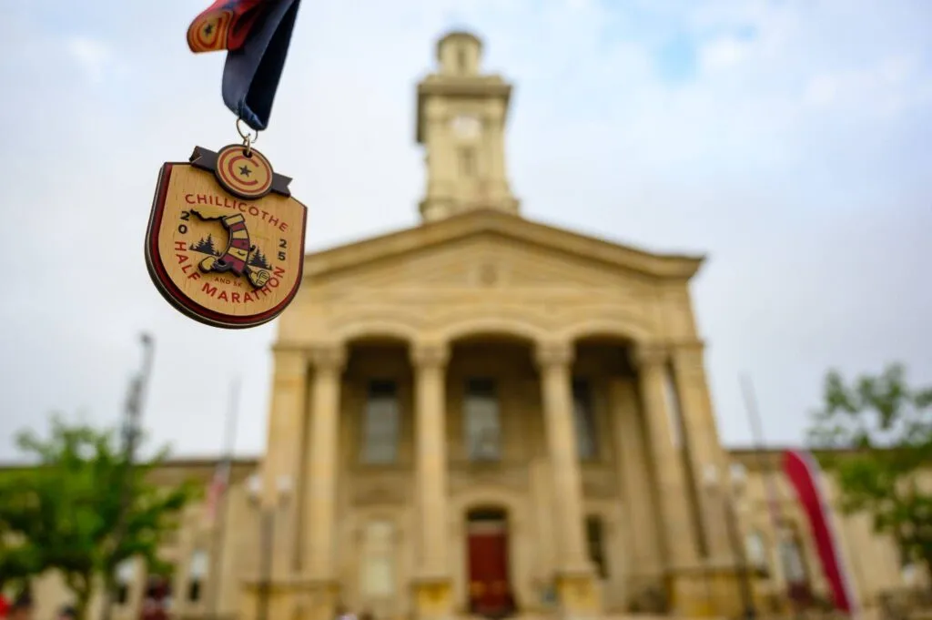 A Chillicothe Half Marathon medal hangs in the foreground, with a blurred image of a large, classical-style building in the background.