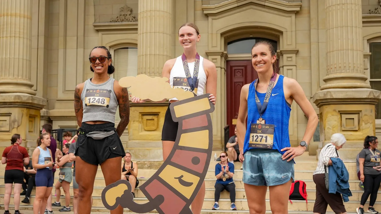 Three female runners smiling on a podium, holding a race-related cutout, with medals around their necks.