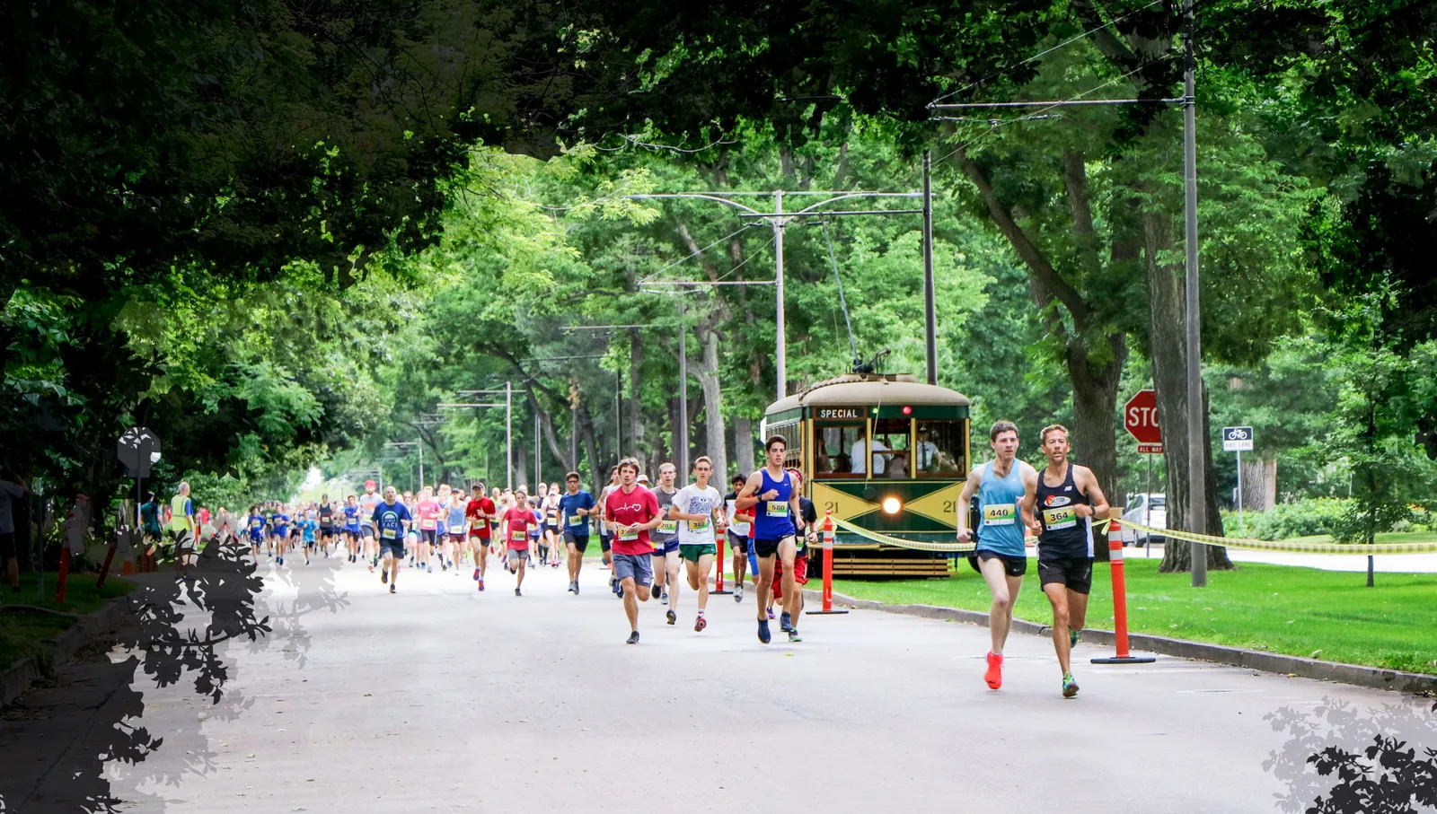 Runners participating in a race on a tree-lined road, with a vintage tram in the background.