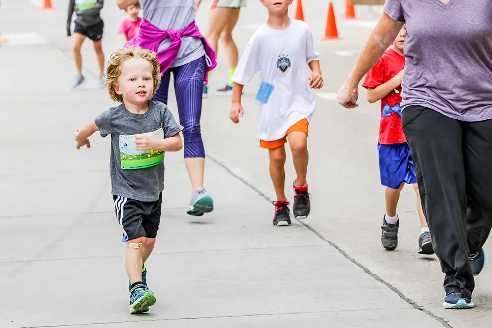 A young boy with blond curly hair runs towards the camera, wearing a grey t-shirt with a race bib and black shorts. He has a bandage on his knee.