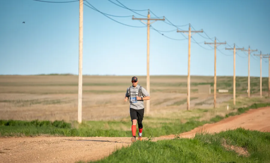 A male runner with bib number 848 runs on a dirt road with telephone poles in the background.