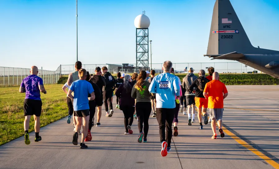 Runners in a race on an airport tarmac, with a large military plane in the foreground and a radar tower in the background.