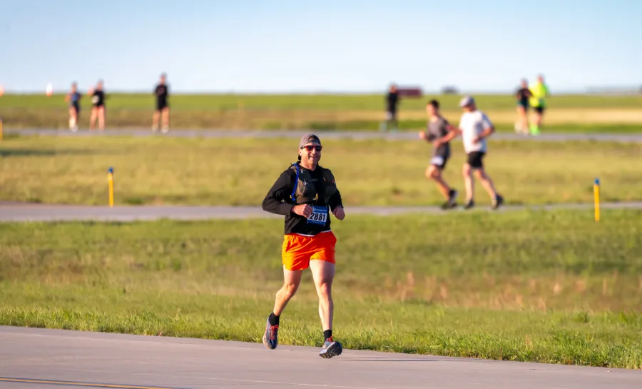 A male runner wearing orange shorts and a black long-sleeved shirt smiles as he runs on a paved path with grass on either side. Other runners are visible in the blurred background.