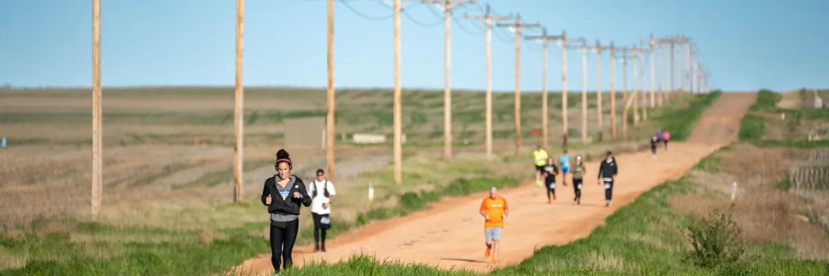 Runners on a dirt road with telephone poles stretching into the distance under a clear blue sky.