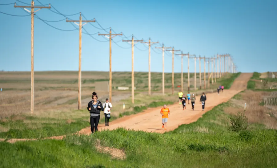 Runners on a dirt road with telephone poles stretching into the distance under a clear blue sky.