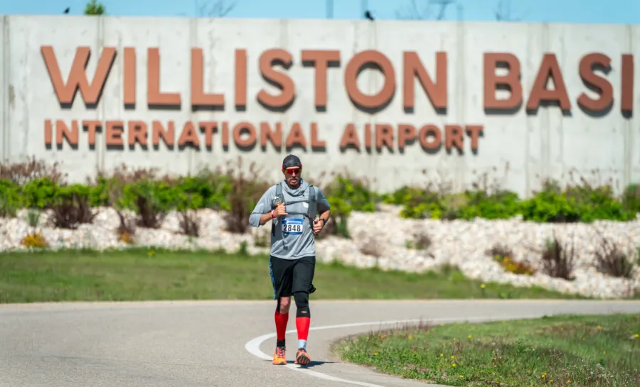 A male runner wearing a grey shirt, black shorts, and red compression socks runs towards the camera in front of a large concrete sign that reads 'WILLISTON BASIN INTERNATIONAL AIRPORT'.