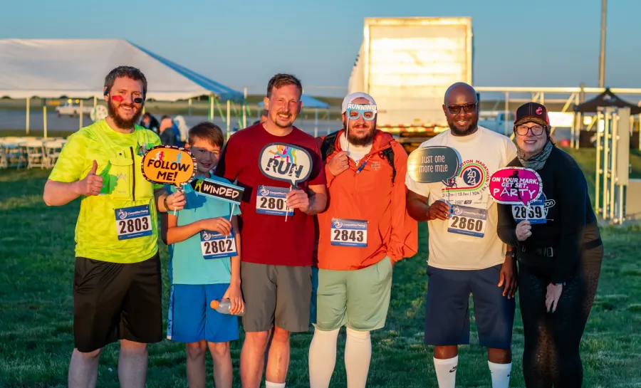 A group of six people, including a child, posing for a photo before a race, holding fun signs and wearing race bibs.