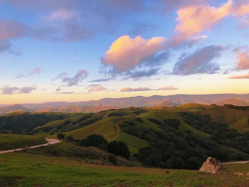 A winding road through rolling green hills at sunset, with colorful clouds in the sky.