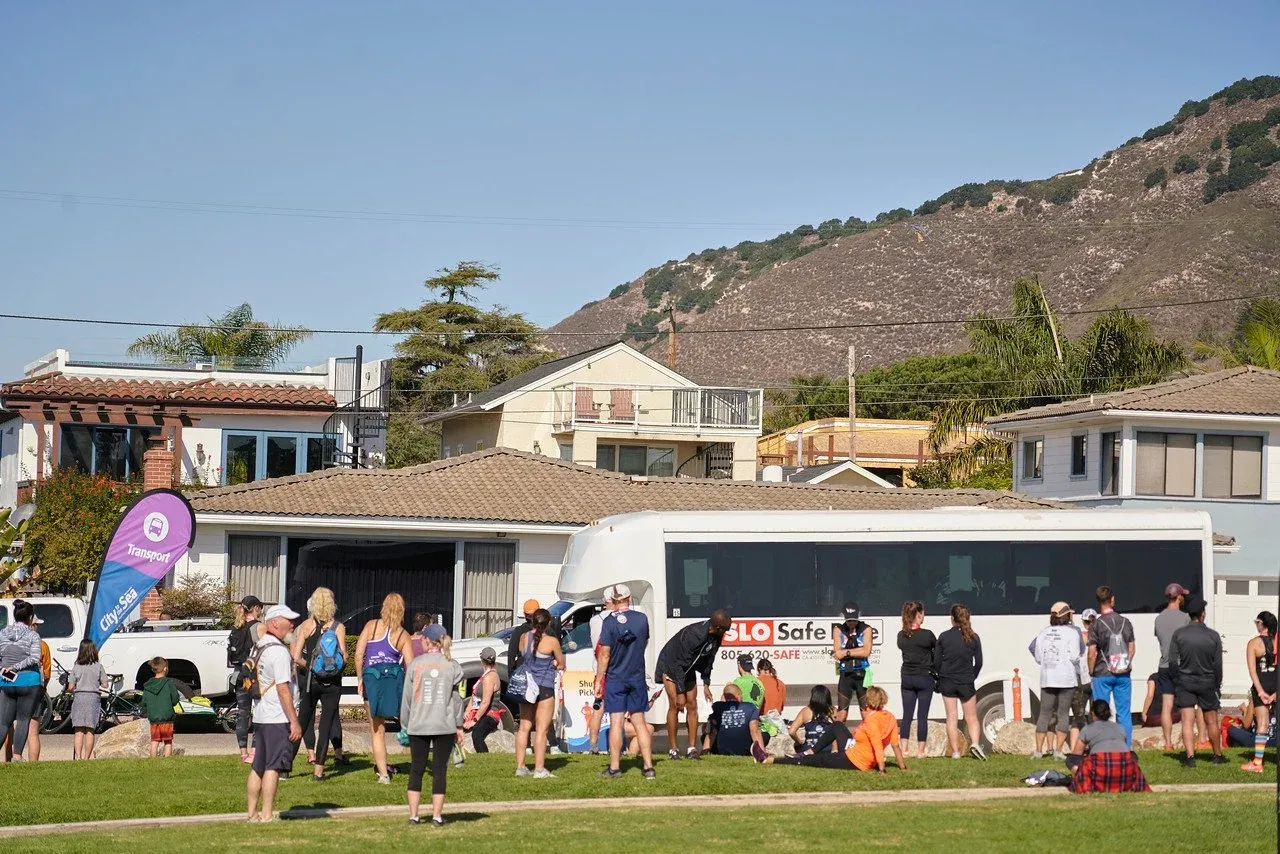 A group of runners and spectators gathered on a grassy area with houses and a bus in the background.