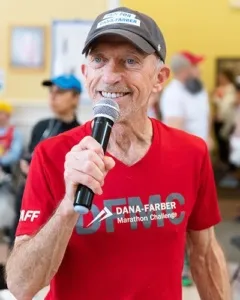 Man in a red Dana-Farber Marathon Challenge shirt and a baseball cap speaking into a microphone.