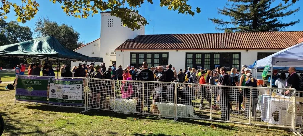 A crowd of people gathered behind a fence at an outdoor event, with tents and buildings in the background.