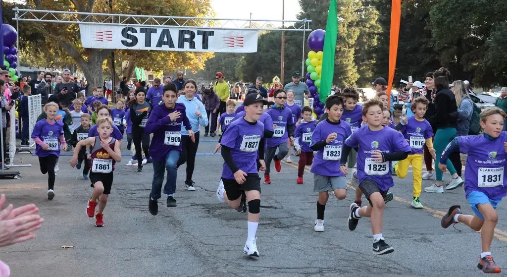 A group of young runners in purple shirts and bib numbers burst forward from the start line, with a banner overhead reading 'START'.