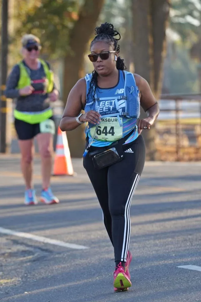A female runner wearing sunglasses and a blue running vest with bib number 644, running on a paved road during a race.
