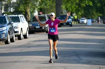 A female runner with bib number 150 smiles and waves with both hands raised as she runs down a paved road lined with parked cars and trees.