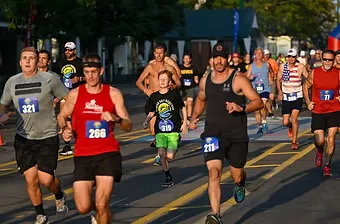 A group of male runners in racing bibs are running down a paved road, with spectators and buildings visible in the background.