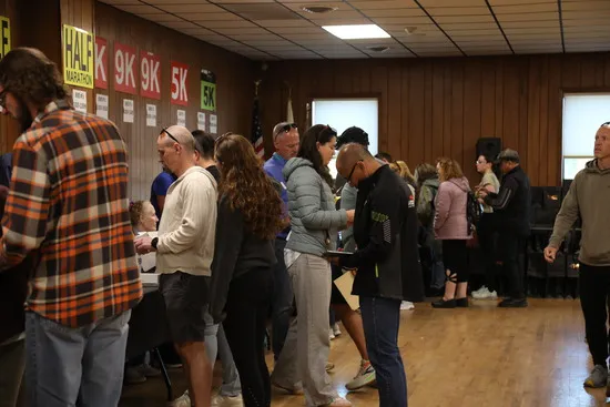 People lining up indoors, with race distance signs like 'HALF MARATHON', '9K', and '5K' visible on the wall.