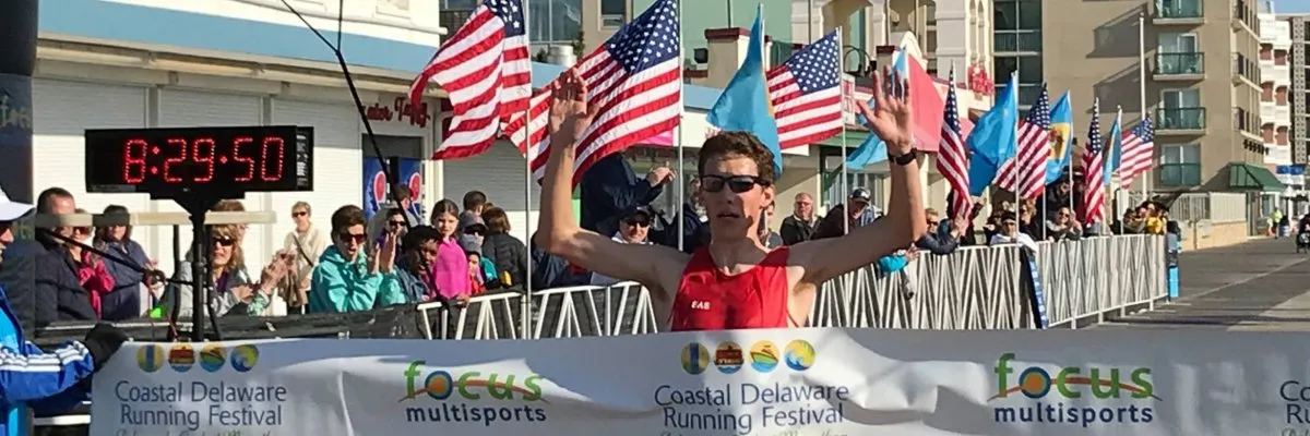 A runner crosses the finish line with arms raised in celebration, with spectators and American flags in the background.