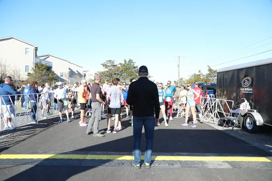 A crowd of runners waits at the start line of a road race on a sunny day.