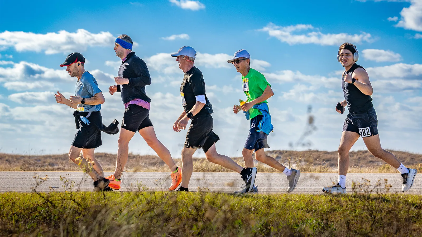 A group of male runners race along a paved road with a grassy shoulder and a bright blue sky with clouds.