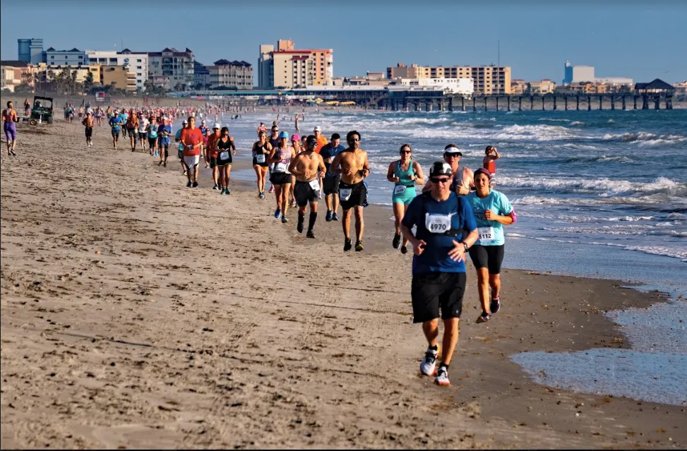 Runners race along a sandy beach with the ocean on their right and buildings in the background.