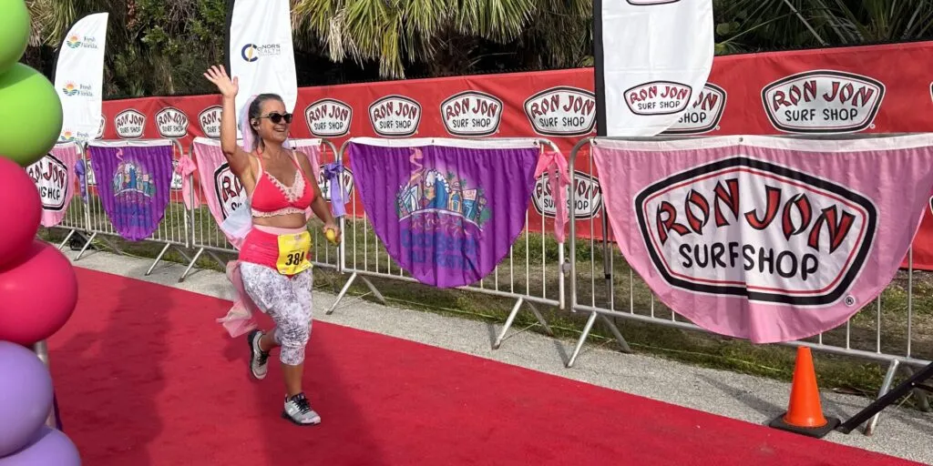 A female runner waves to the camera while running on a red carpet, with sponsor banners in the background.