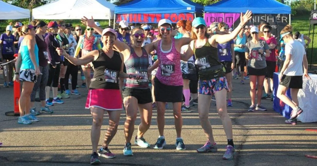 A group of female runners smiling and posing at the start line of a race, with tents and other participants in the background.