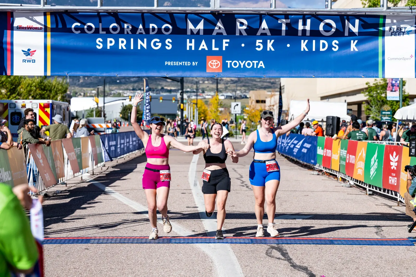 Three female runners, two holding hands, cross the finish line under a Colorado Springs Marathon banner, smiling and celebrating.