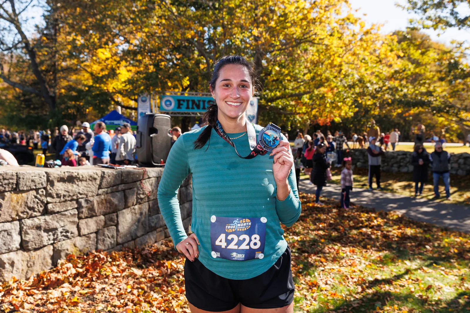 A smiling female runner holds up her race medal in front of a finish line banner, with autumn trees and a crowd in the background.