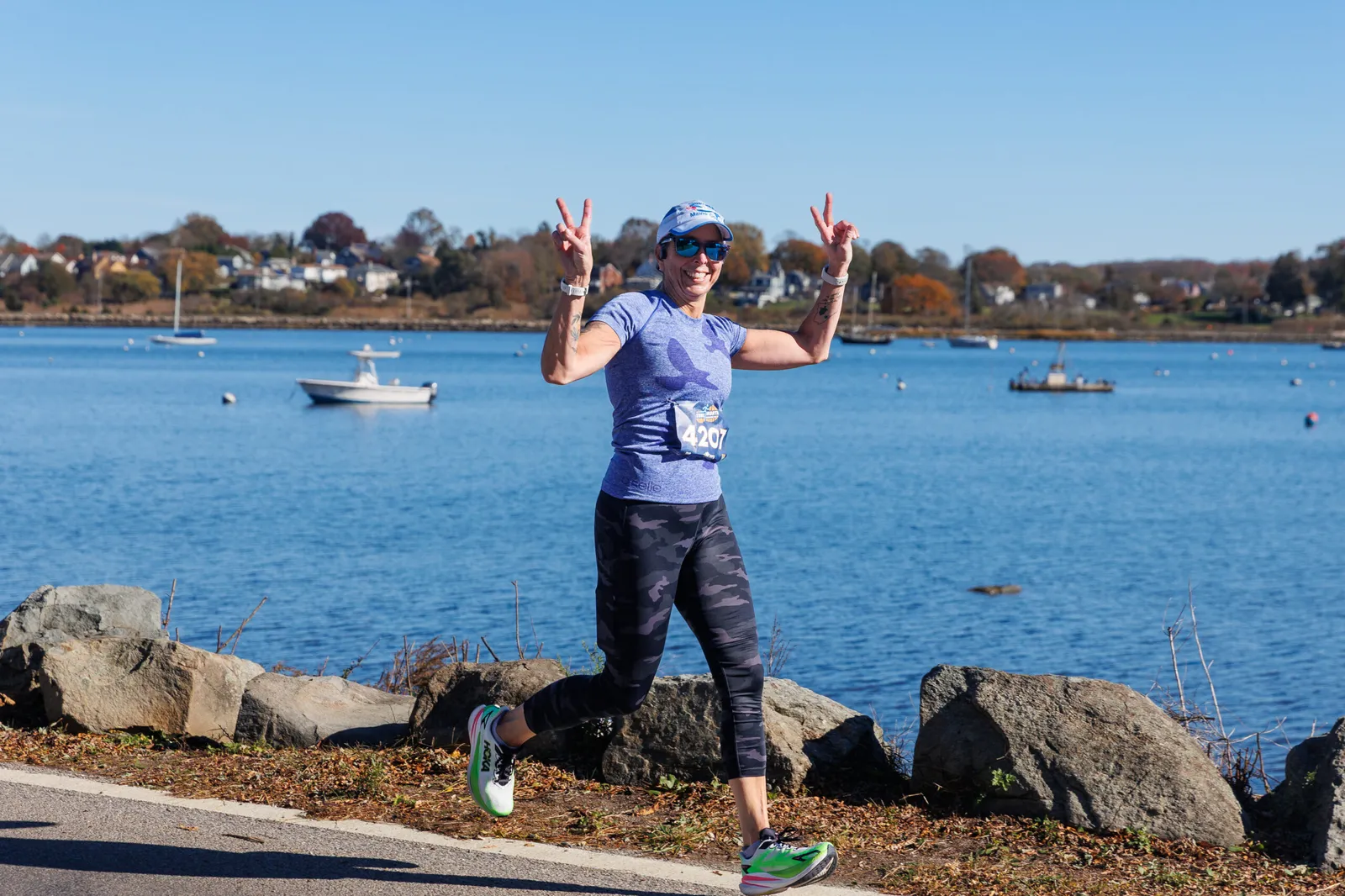 A female runner smiles and holds up two peace signs while running along a waterfront path.