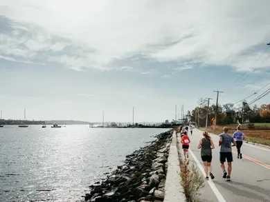 Runners on a road next to a body of water with boats in the distance.