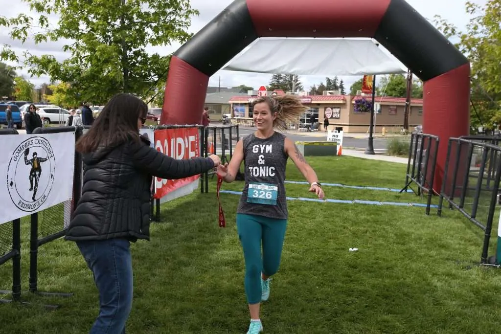 A female runner with a bib number 326 crosses the finish line under an inflatable arch, smiling.