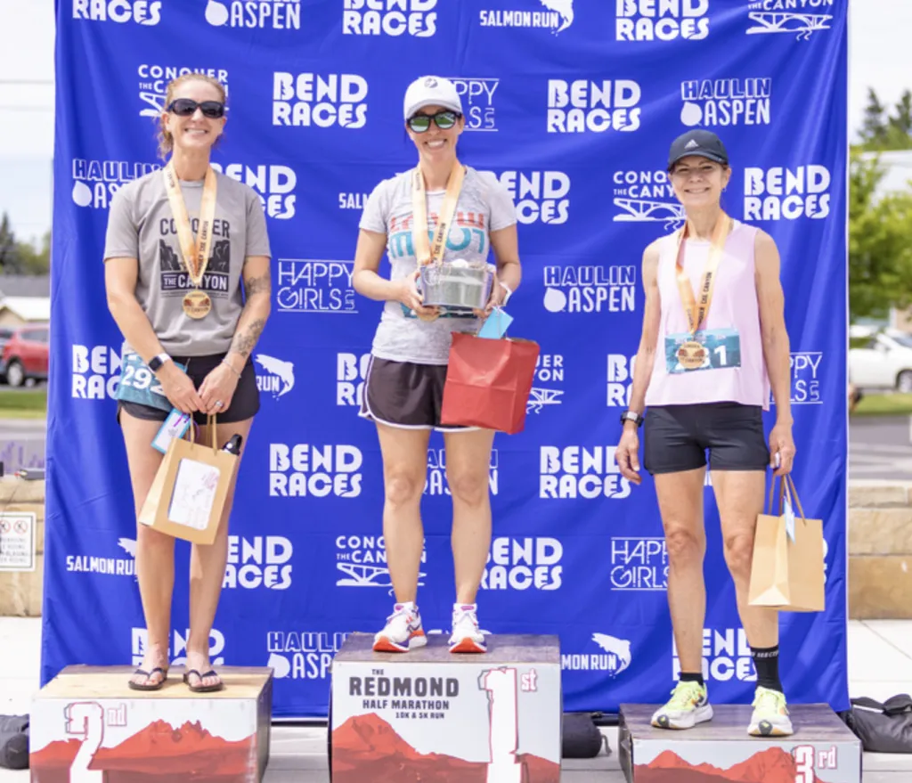 Three female runners on a podium, smiling and holding awards, with a blue banner in the background displaying race sponsor logos.