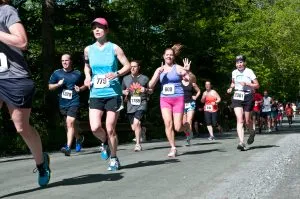 A group of runners on a paved road, with trees and foliage in the background. One runner in the foreground is wearing a blue tank top and black shorts, and another runner in a purple tank top and pink shorts is waving.