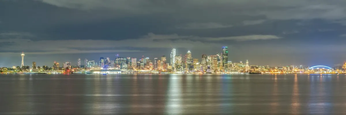 Panoramic nighttime view of the Seattle skyline with the Space Needle and waterfront lights reflected in the water.