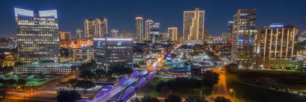 Nighttime cityscape with illuminated skyscrapers and a bridge with purple lights.