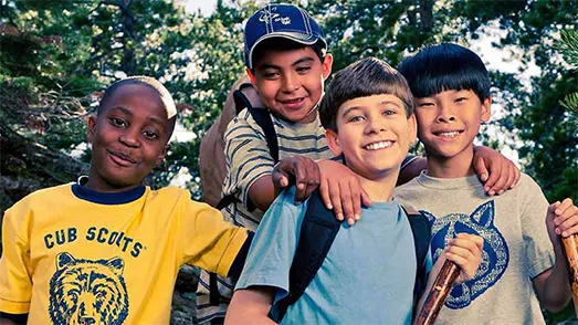 Four smiling young boys with backpacks and walking sticks posing for a photo in a forest setting.