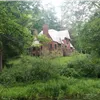 A rustic stone building with a large chimney sits on a grassy hill surrounded by lush green trees.
