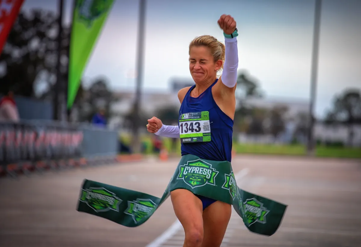 A female runner with her arm raised in triumph as she crosses the finish line, breaking through a green banner with the race logo.
