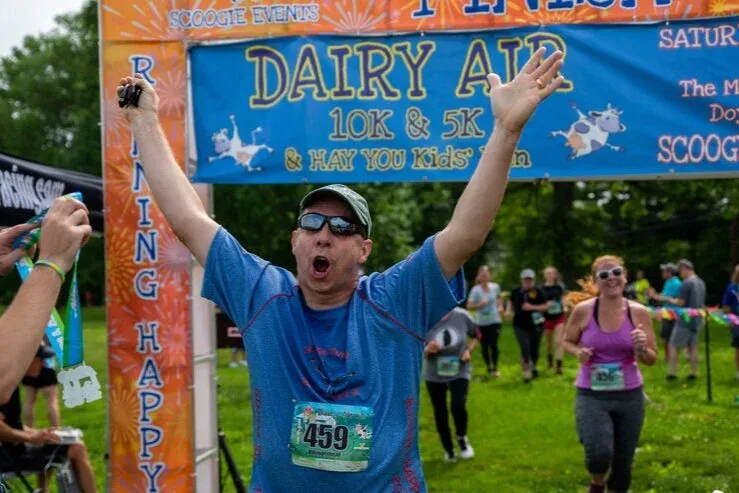 A male runner with his arms raised in celebration crosses the finish line at the Dairy Aid 10K & 5K race, with other runners and the race banner in the background.