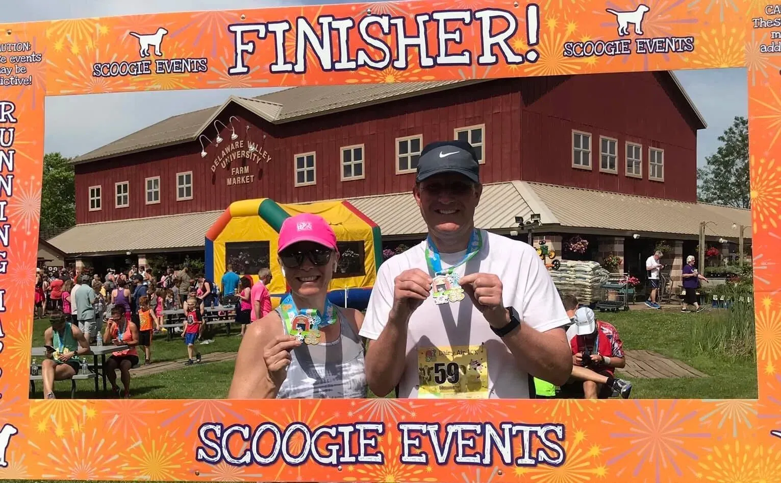 A smiling couple holds up their race medals in front of a red barn and a 'FINISHER!' banner.