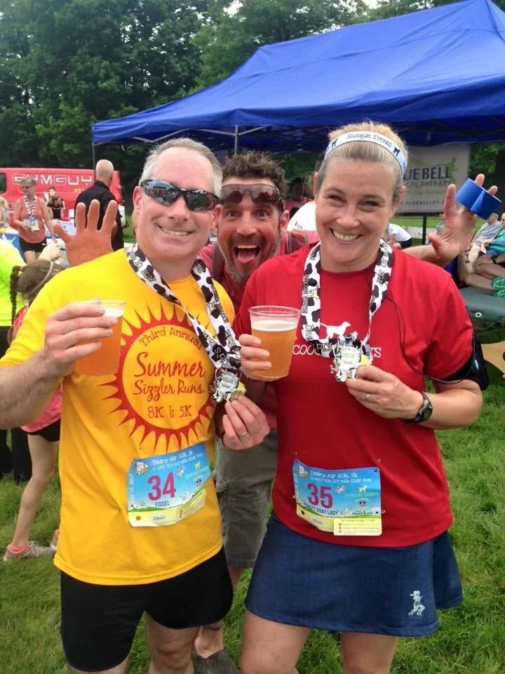 Three smiling runners, two wearing medals and holding drinks, pose for a photo after a race.