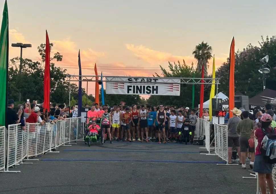 Runners line up at the start/finish line under a banner, with spectators watching and colorful flags lining the course, during sunset.