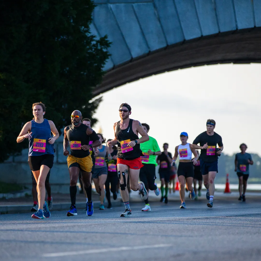A group of runners in a half marathon race, with several runners in the foreground and more in the background, running under a large bridge structure.