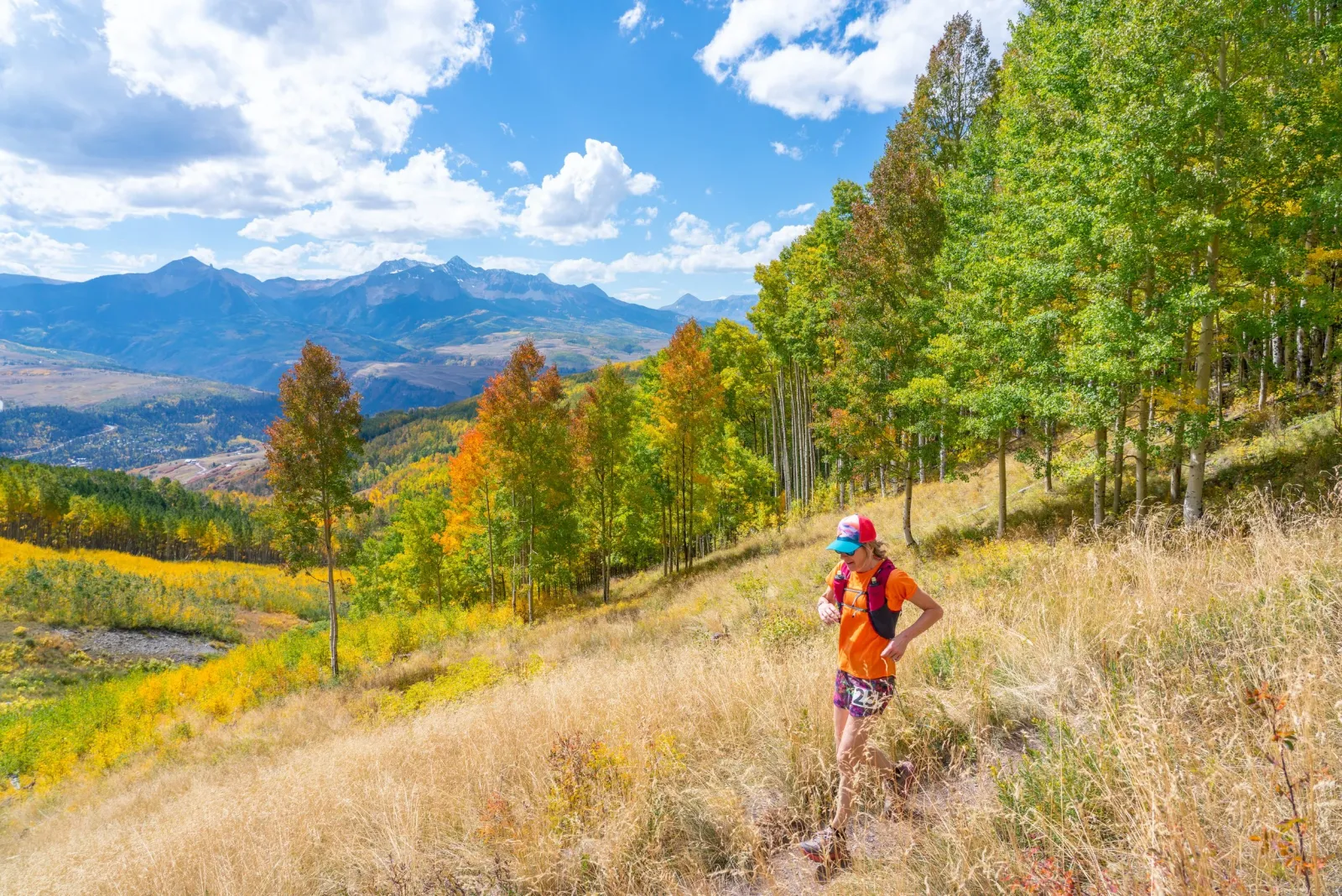 A solo runner on a scenic mountain trail with colorful autumn foliage and distant mountains under a blue sky with clouds.