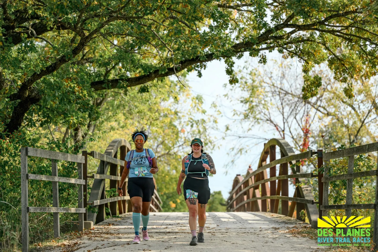 Two female runners smile as they run across a wooden bridge on a tree-lined trail.