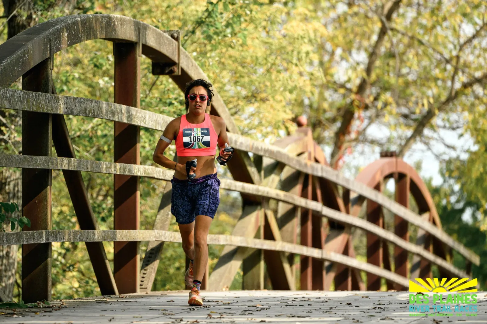 A female runner wearing sunglasses and a pink tank top with bib number 1067 runs across a wooden bridge on a trail.