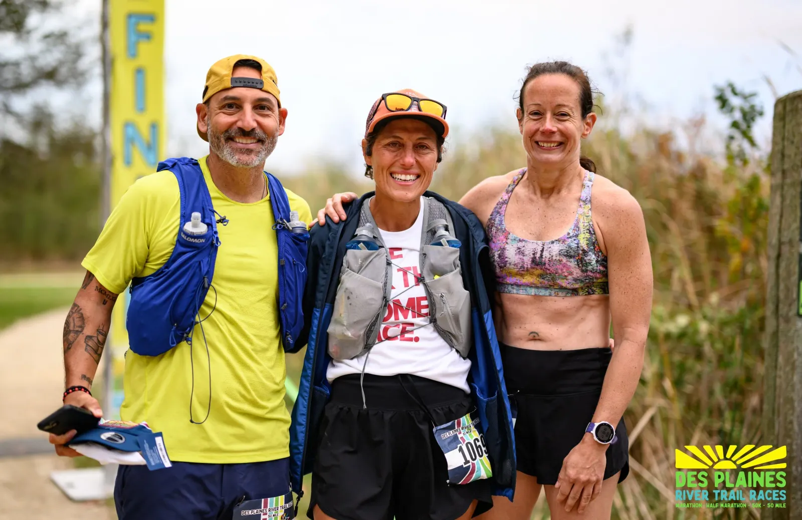 Three smiling runners pose for a photo near a finish line banner at the Des Plaines River Trail Races.